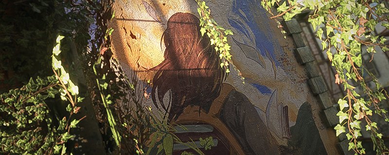 The mural of a figure with long hair walking away from the camera; their hair is long and brown, they carry a backpack full of books and have another book open in front of them. The mural is framed by ivy.